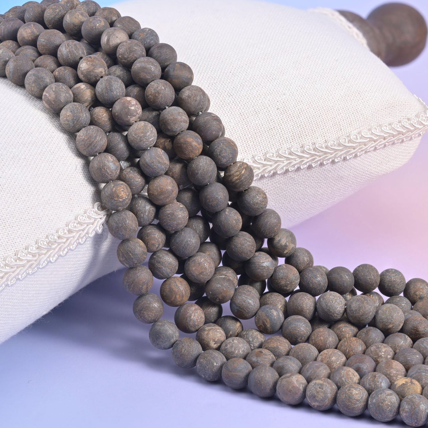 Matte bronzite round bead strands in mixed sizes resting against a white linen cushion, dry chalky surface with no shine