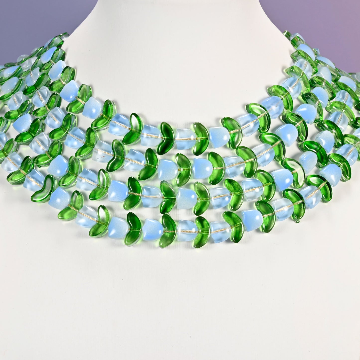 Multi-strand necklace of light blue tulip and green leaf glass beads laid on a white mannequin neck form against a lavender background.