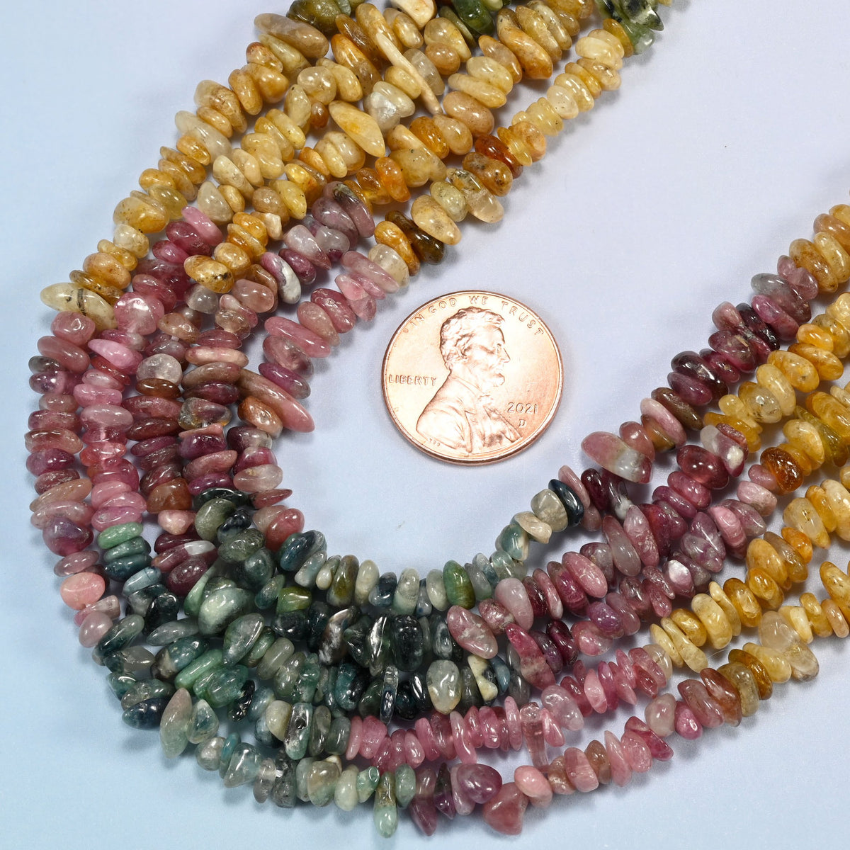 Close view of multicolor tourmaline chip strands curving beside a US penny for scale