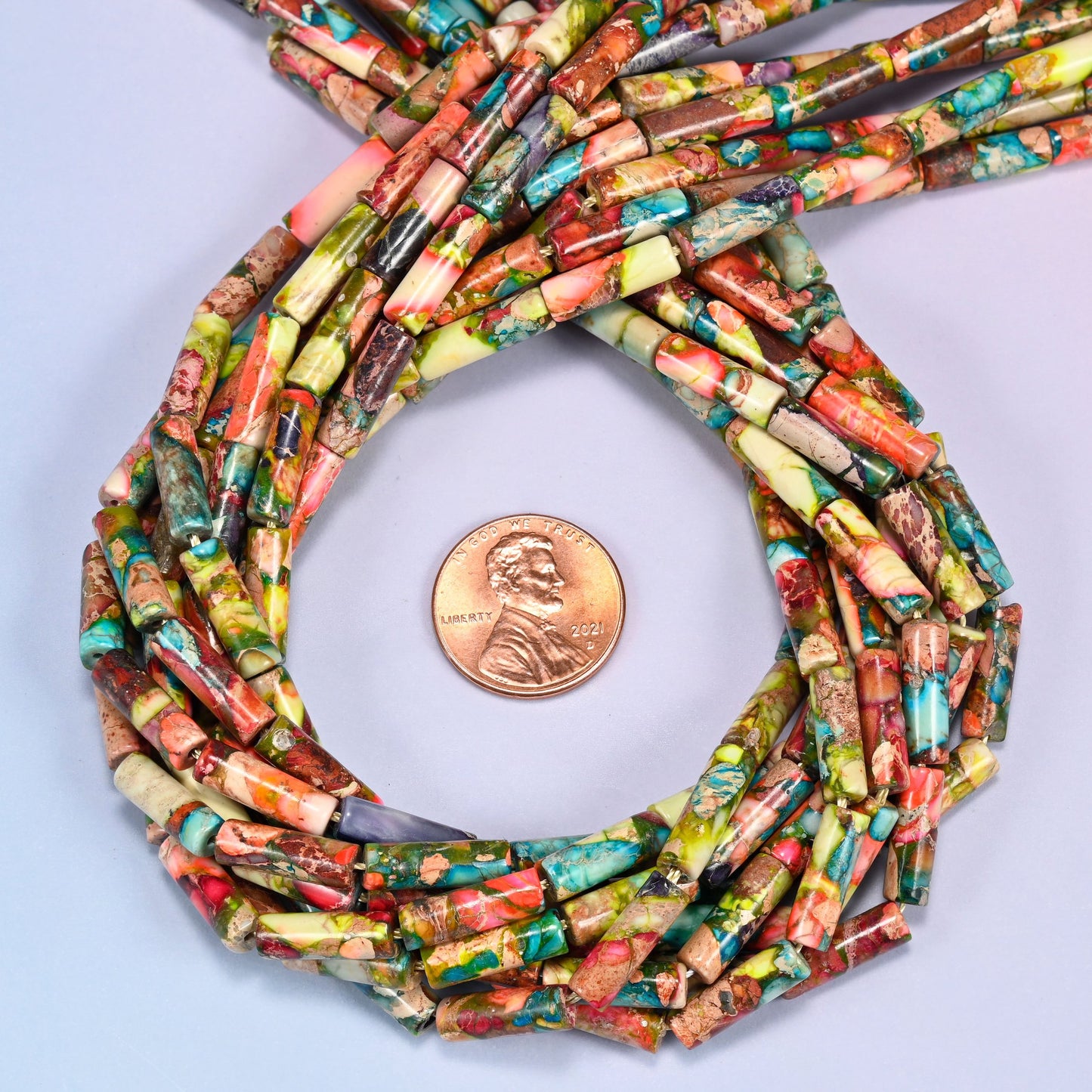 Bundled coil of colorful jasper tube beads on a lavender background with a penny placed at the center for scale.