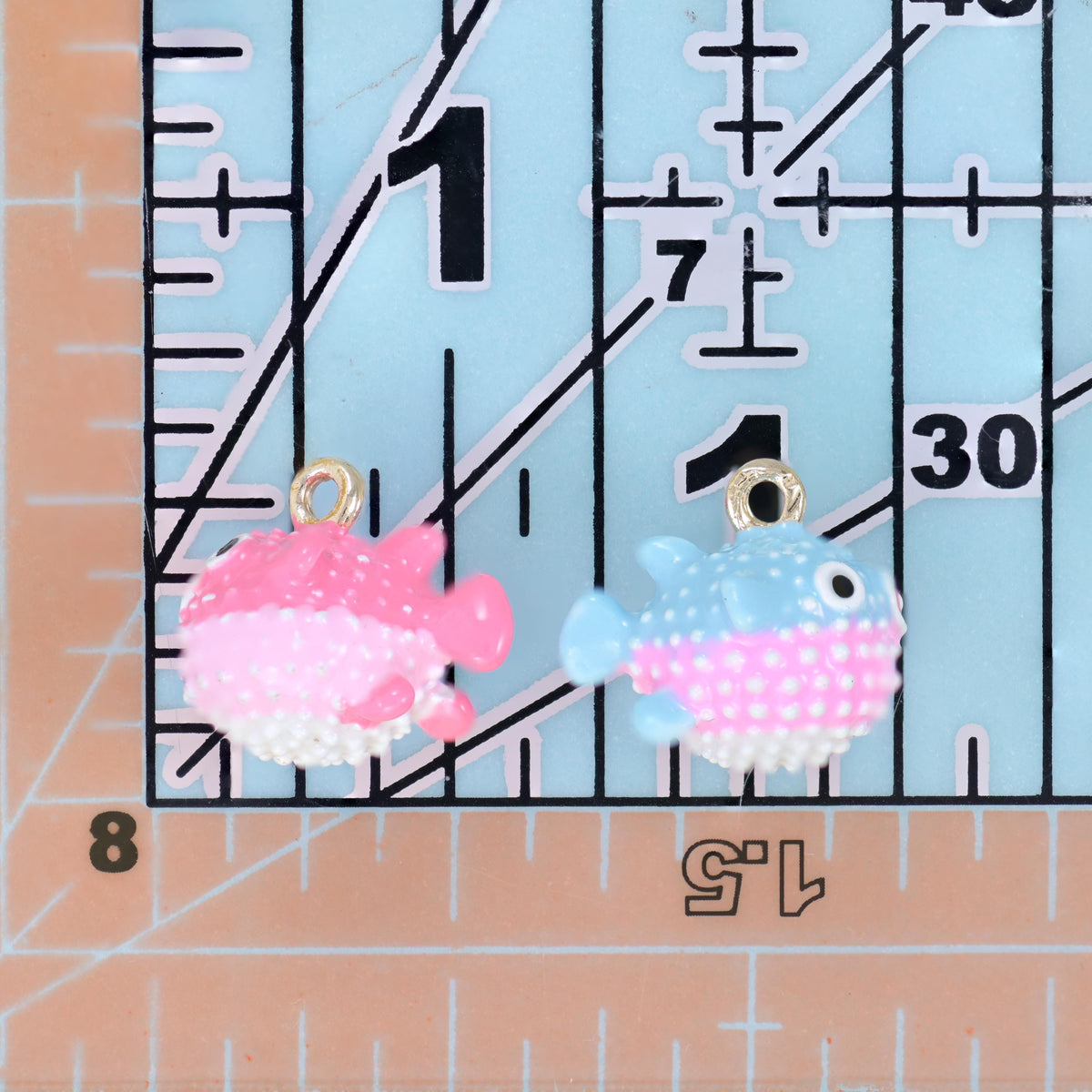 Enamel blowfish charms in pink and light blue placed side by side on a ruler for size comparison.