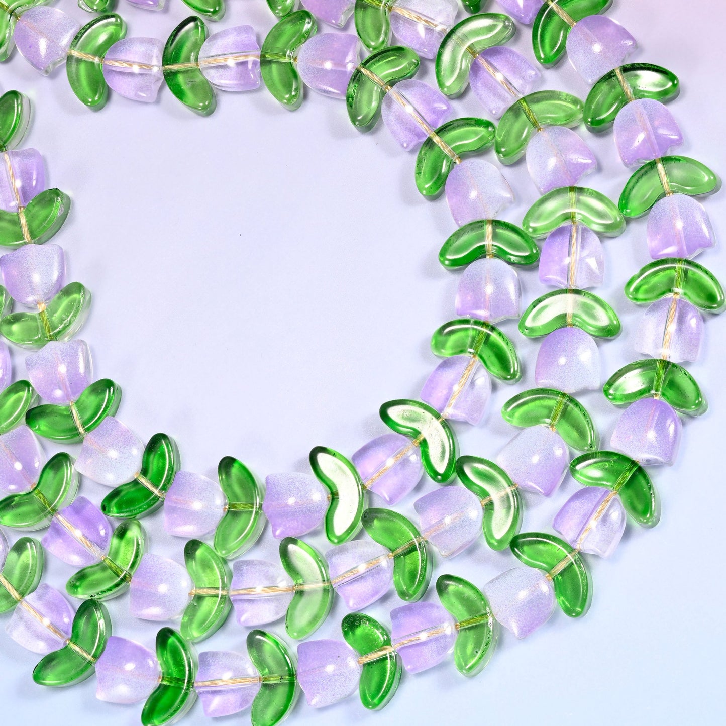 Wide view of a circular coil of lavender tulip and green leaf glass bead strands on a lavender and pink gradient background.