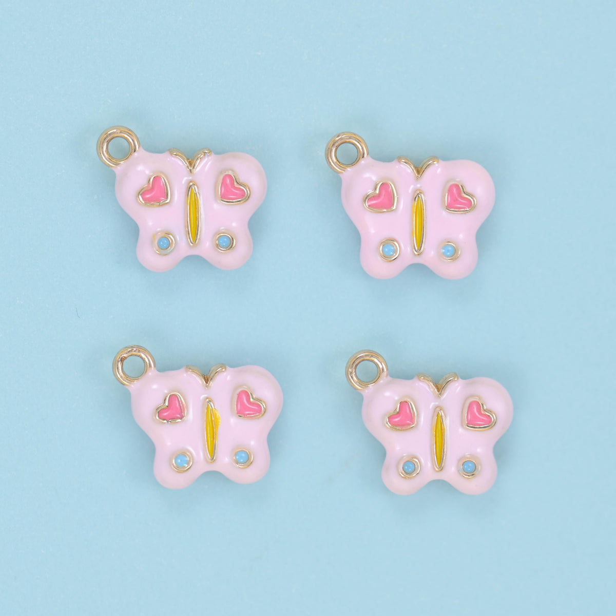 Enamel butterfly charm in soft pink, shown as four charms with deeper pink hearts, blue dots and gold jump rings set above the head.