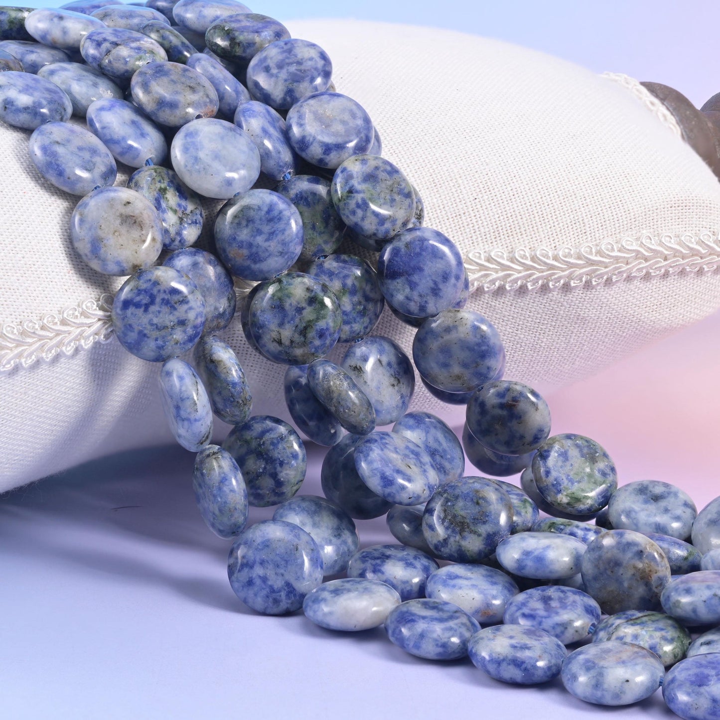 Blue spot jasper coin strands resting on a cream cushion, pale grey discs speckled with powder blue