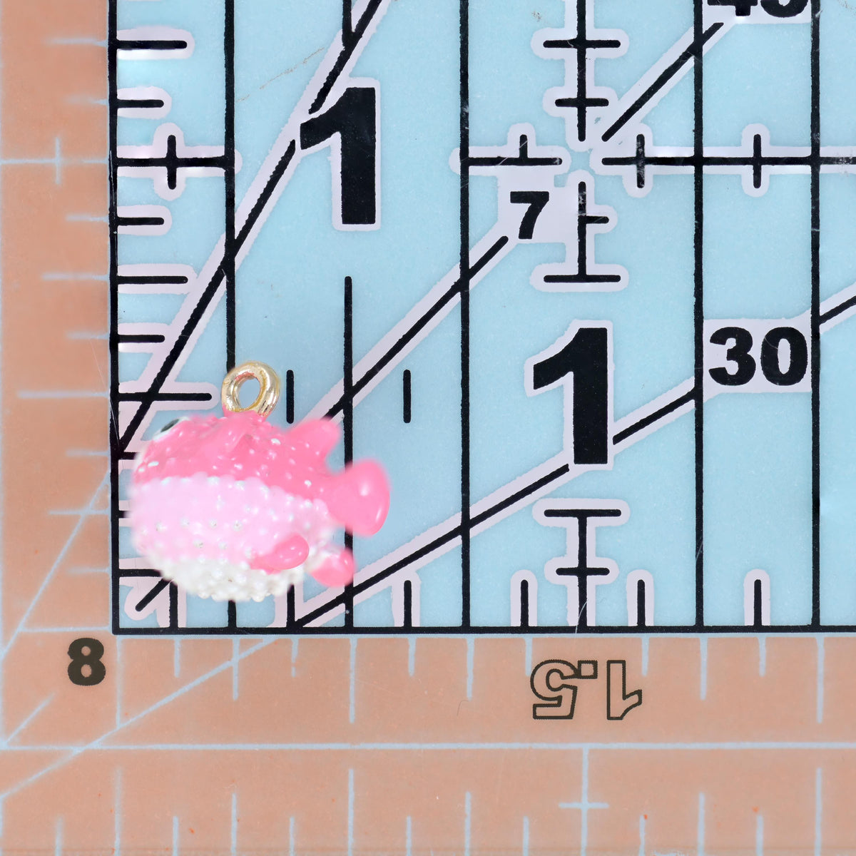 Enamel blowfish charm in pink on a quilting ruler, sitting just under the 1 inch mark for scale.