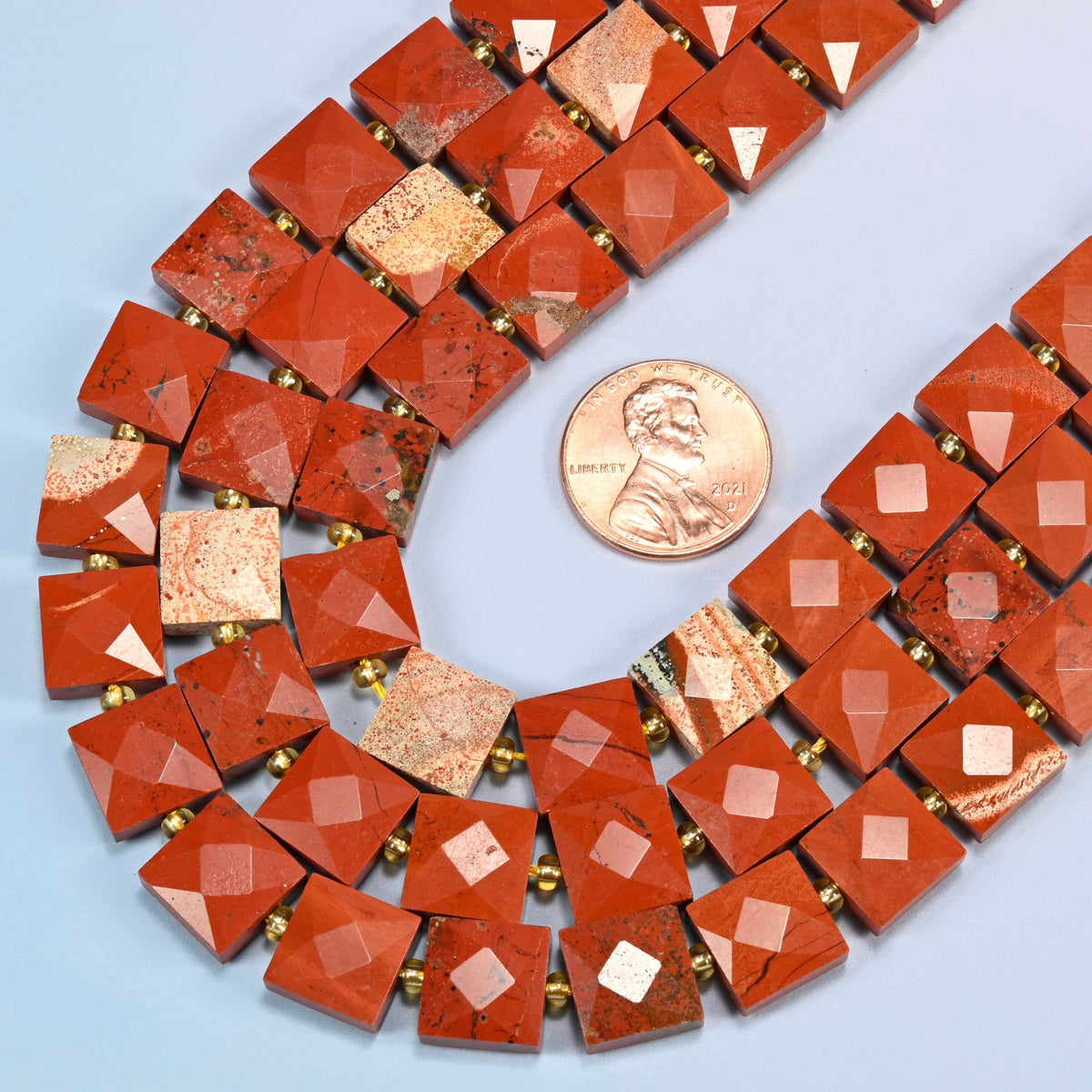 Close view of natural flower red jasper faceted flat square strands curving beside a US penny for scale
