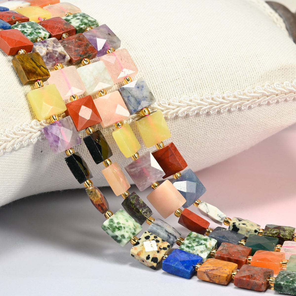 Assorted gemstone faceted flat square beads 10x10mm with small gold seed bead spacers, several strands draped over a white cushion