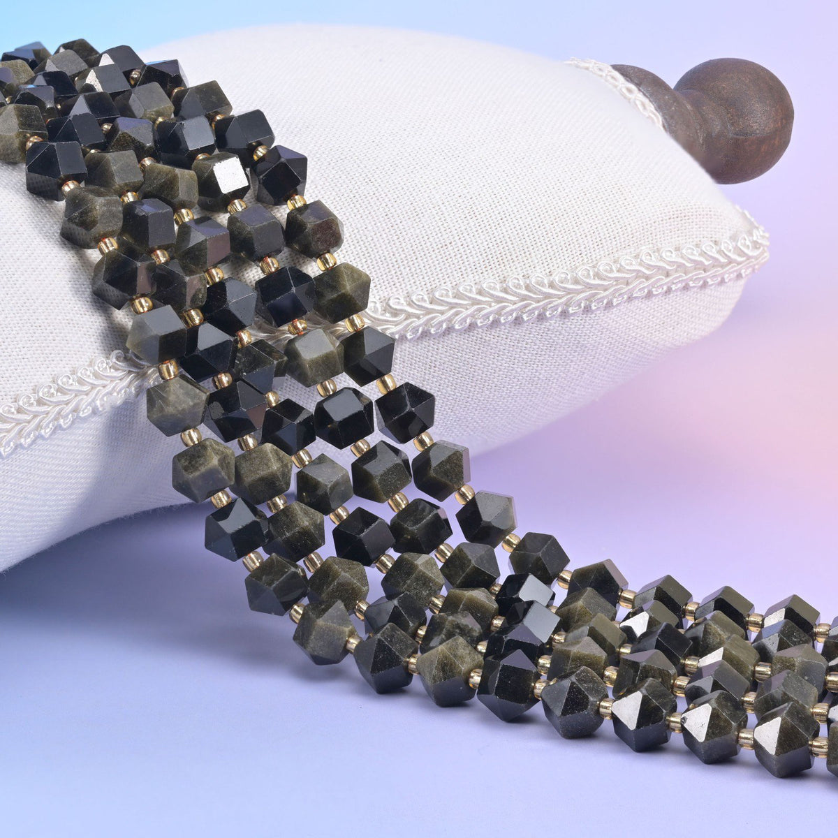 Faceted 8 sides diamond rhombus beads in gold obsidian draped over a white cushion, dark beads with a golden sheen.