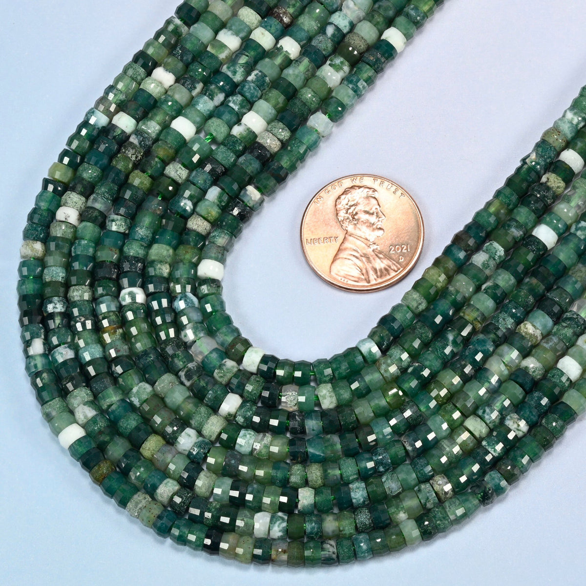 Close view of moss agate faceted heishi disc strands curving beside a US penny for scale