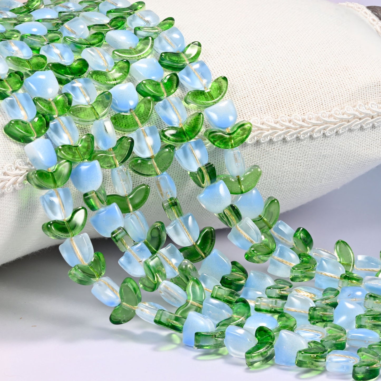 Close-up of light blue opal-toned tulip flower and green leaf glass beads cascading diagonally over a cream woven pillow with decorative trim.