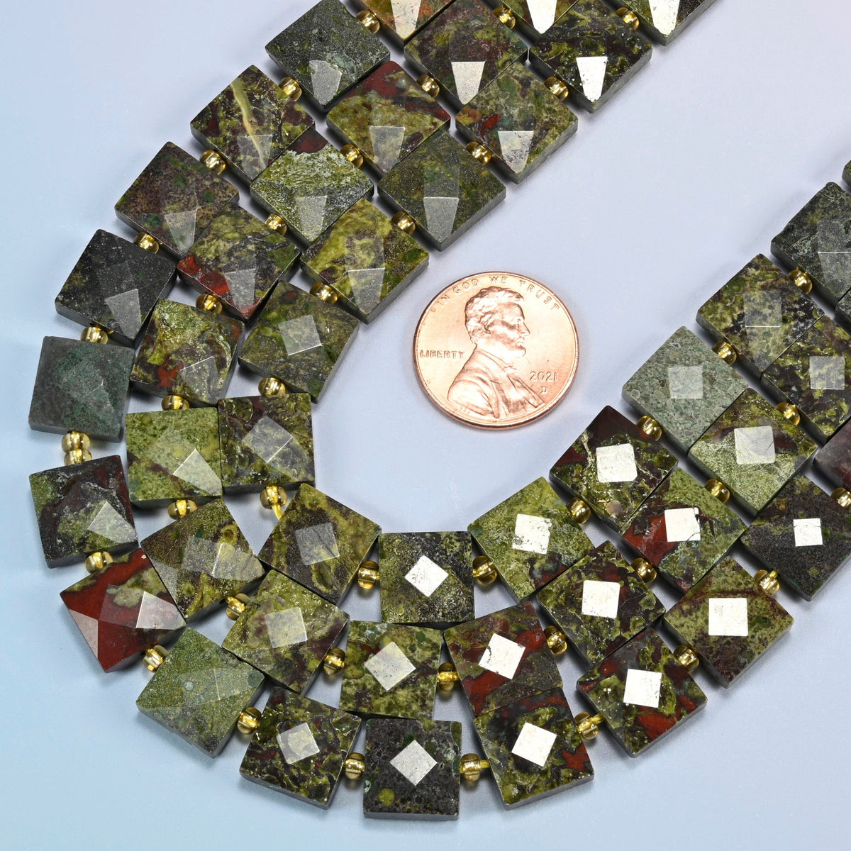 Close view of natural dragon blood stone faceted flat square strands curving beside a US penny for scale