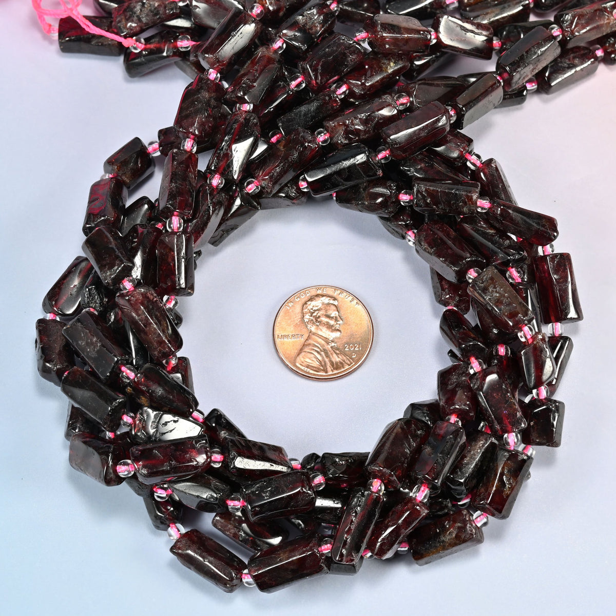 Red garnet tube beads 7x10-7x14mm coiled in a loop around a US penny for scale, close-up of the irregular hand-cut tube shape