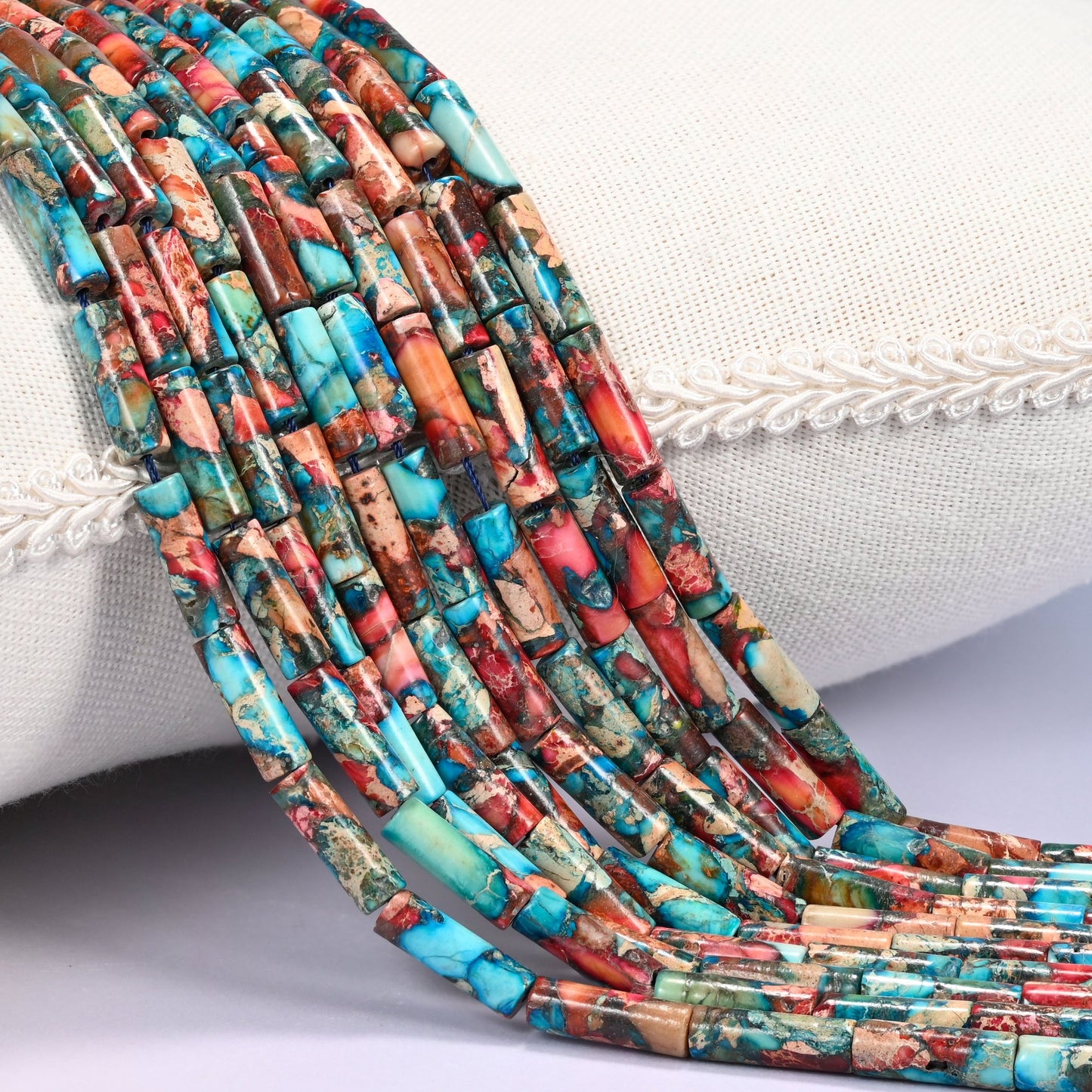 Close-up of multiple strands of turquoise, coral red, and brown dyed Sea Sediment Jasper tube beads draped over a cream fabric jewelry display bust.