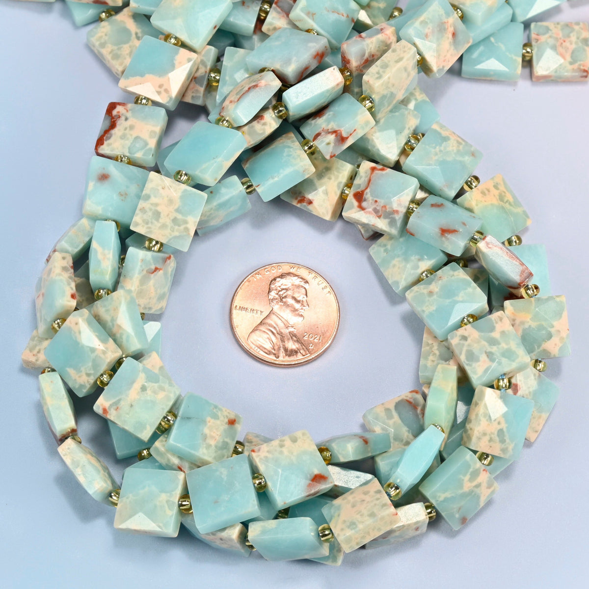 Dyed blue Shoushan stone faceted flat square beads 10x10mm coiled in a loop around a US penny for scale, close-up of the faceted flat square shape