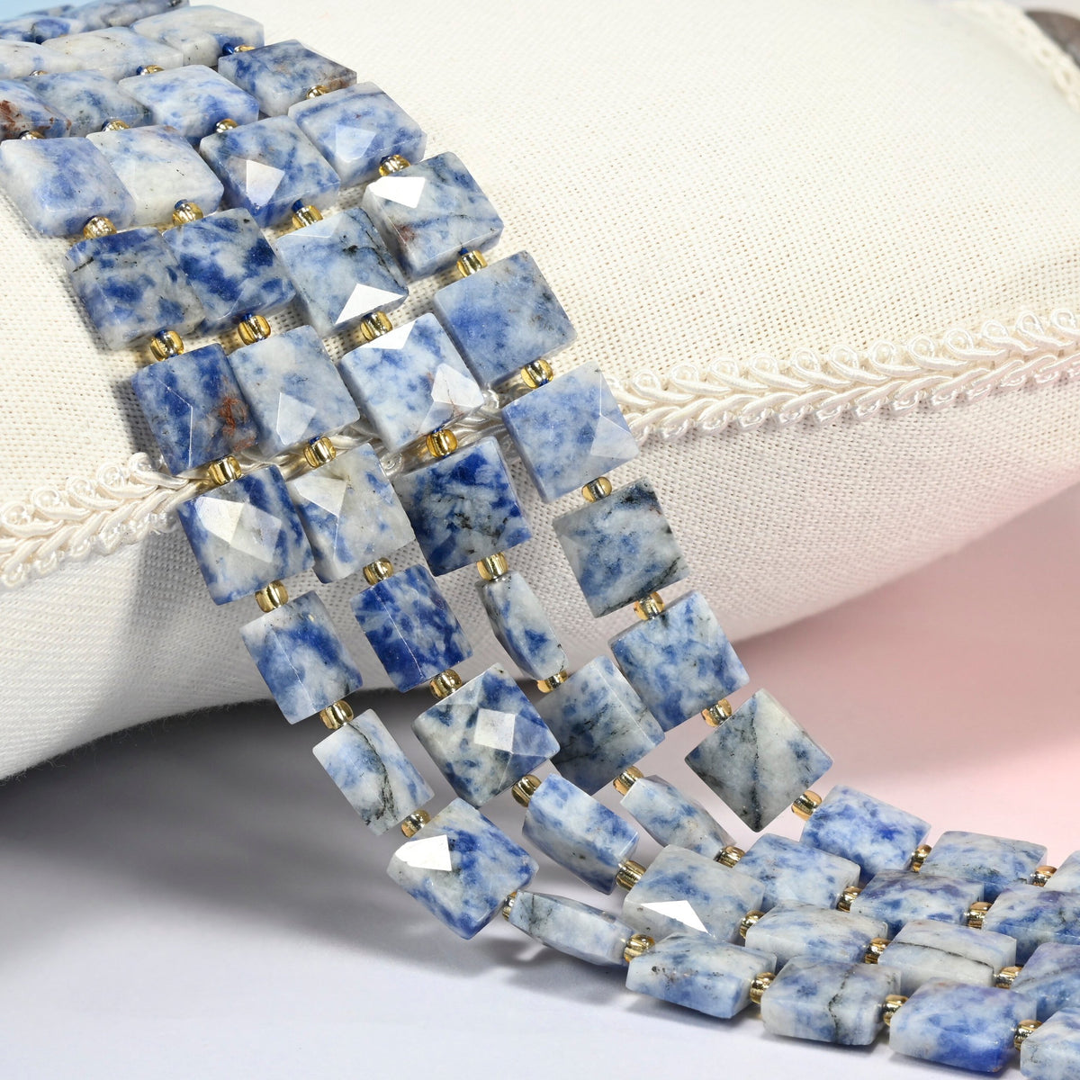 Natural blue spot jasper faceted flat square beads 10x10mm with small gold seed bead spacers, several strands draped over a white cushion