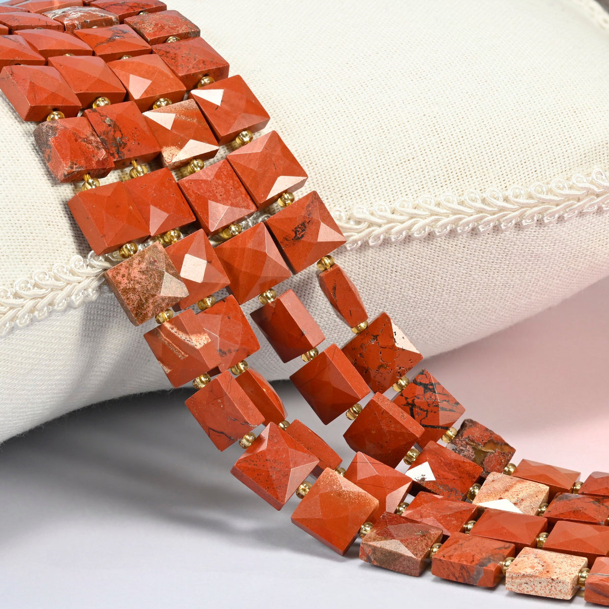 Natural flower red jasper faceted flat square beads 10x10mm with small gold seed bead spacers, several strands draped over a white cushion