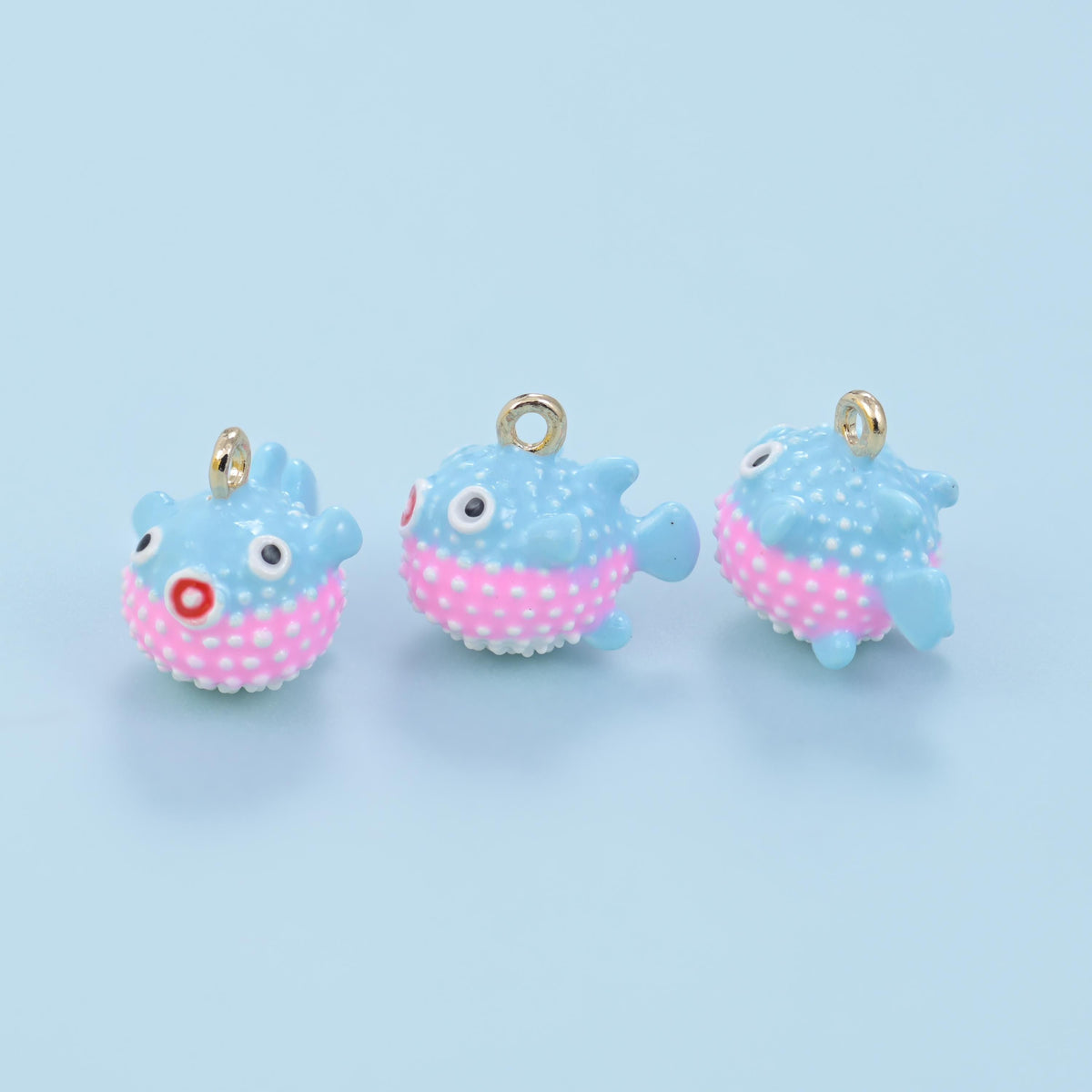 Enamel blowfish charm in light blue with a pink dotted belly, three views on a blue background.