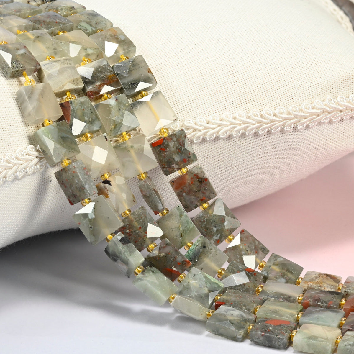 Natural African bloodstone faceted flat square beads 10x10mm with small gold seed bead spacers, several strands draped over a white cushion