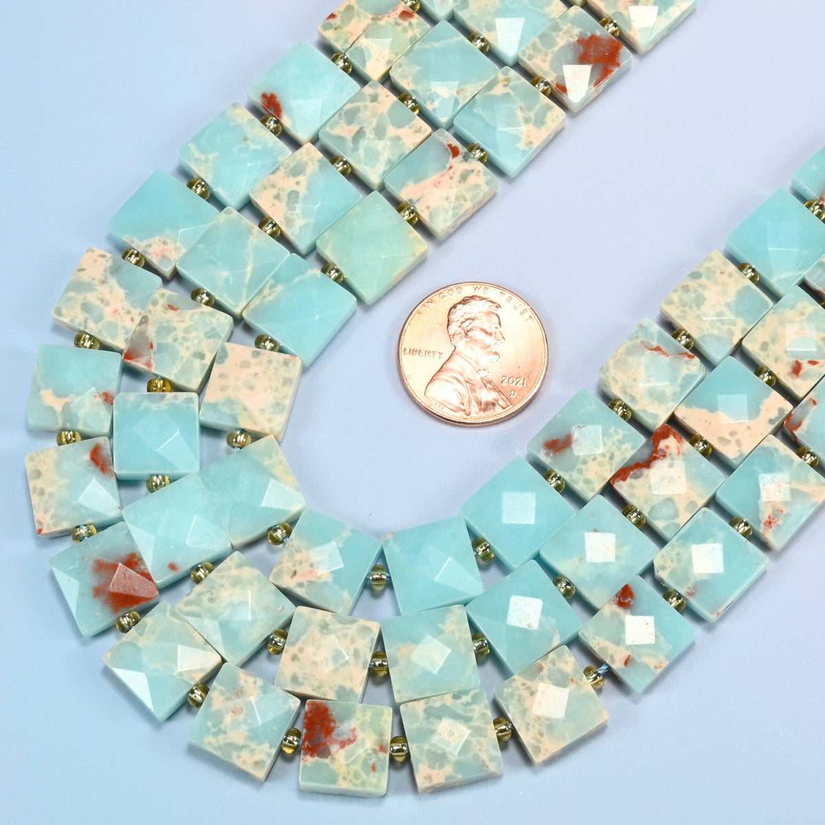 Close view of dyed blue Shoushan stone faceted flat square strands curving beside a US penny for scale
