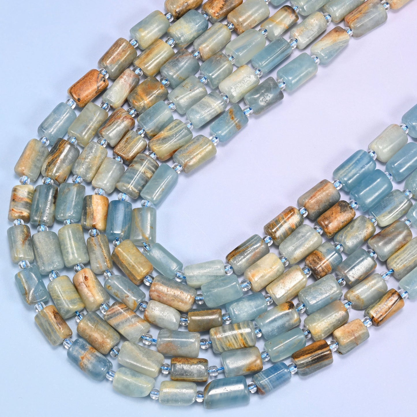 Close-up of faceted Blue Calcite tube beads with clear glass spacer beads on a lavender background, showing natural banding.