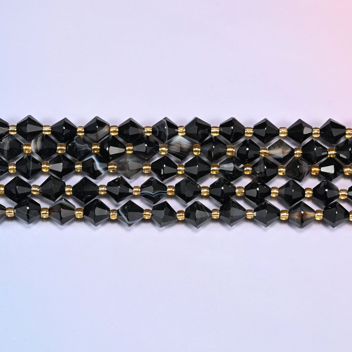Dyed black strip agate bicone beads in straight rows, seen close up, each bead cut to two points meeting at a wide center, facets catching hard highlights.