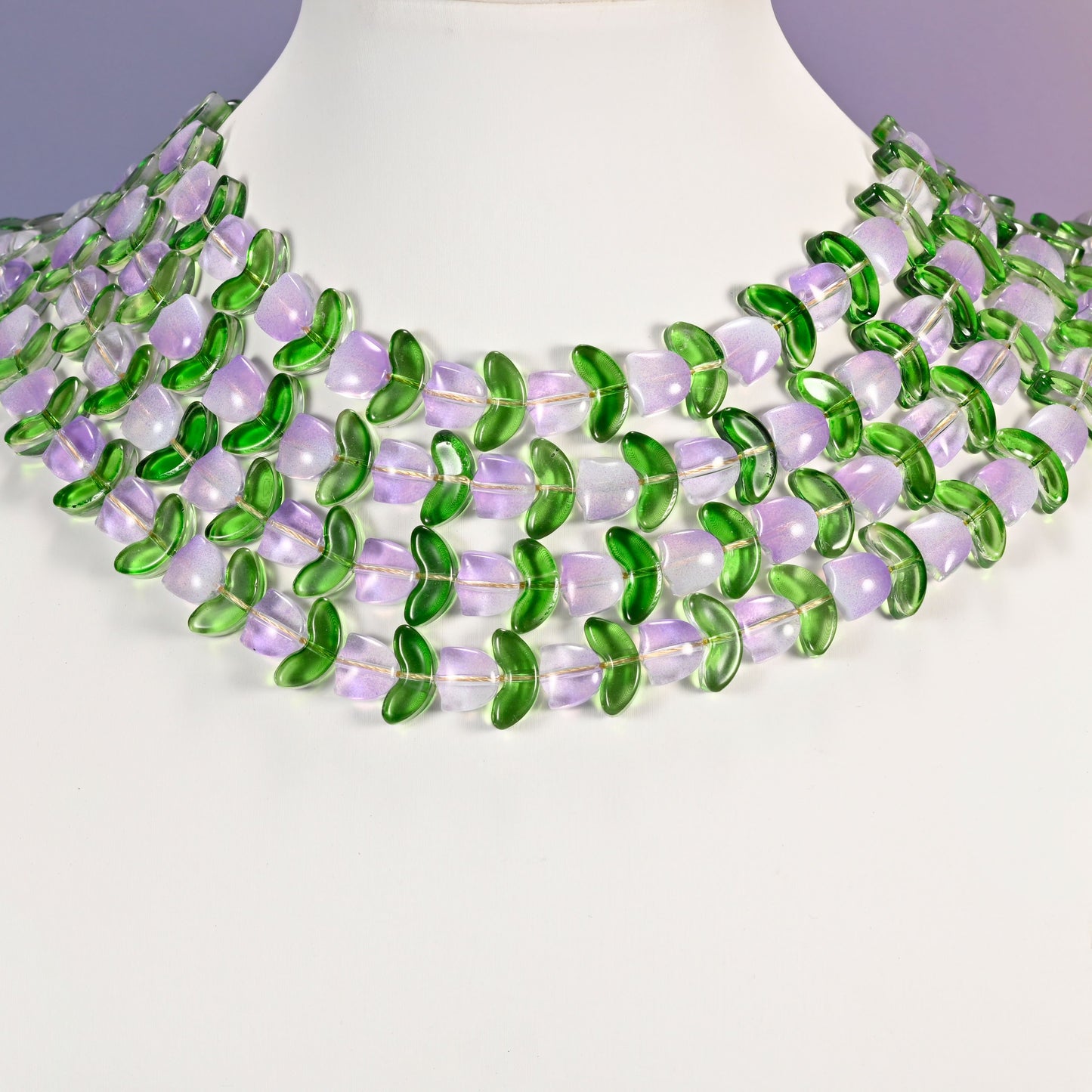 Multi-strand necklace of lavender tulip and green leaf glass beads laid on a white mannequin neck form against a lavender background.