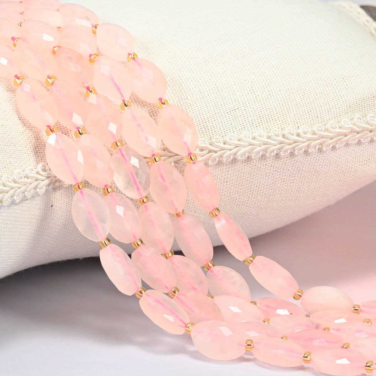 Rose quartz faceted flat oval beads 9x14mm with small gold seed bead spacers, several strands draped over a white cushion