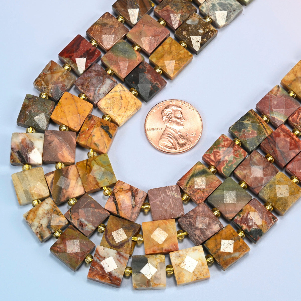Close view of natural red howlite faceted flat square strands curving beside a US penny for scale