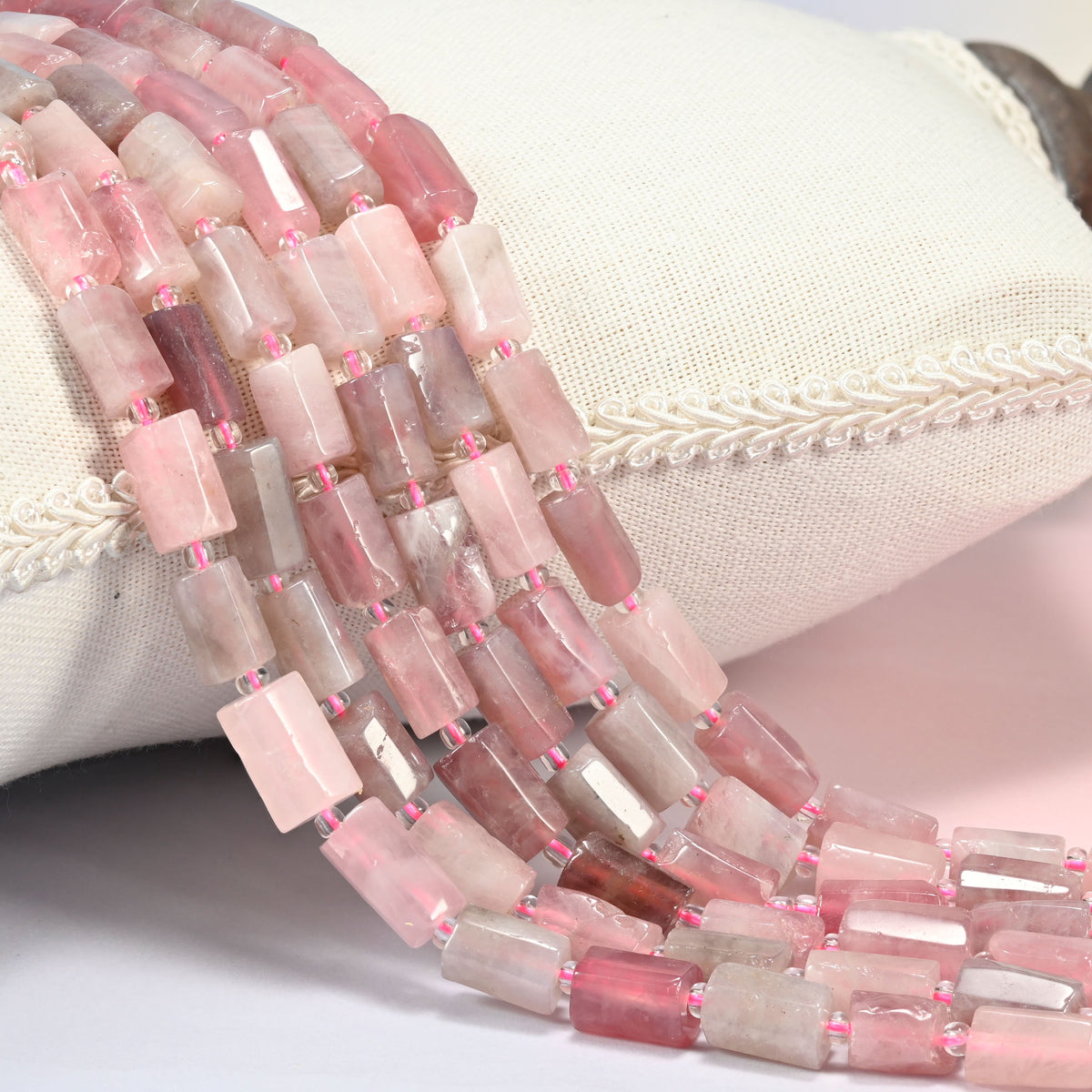 Madagascar multi color rose quartz faceted tube beads draped over a white linen cushion with braided trim, blush and rose pink.