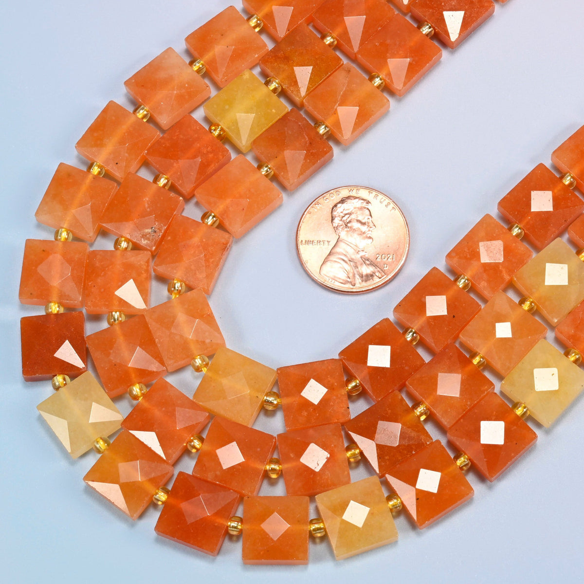 Close view of natural red aventurine faceted flat square strands curving beside a US penny for scale