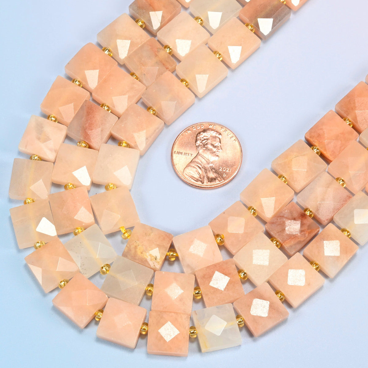 Close view of natural pink aventurine faceted flat square strands curving beside a US penny for scale