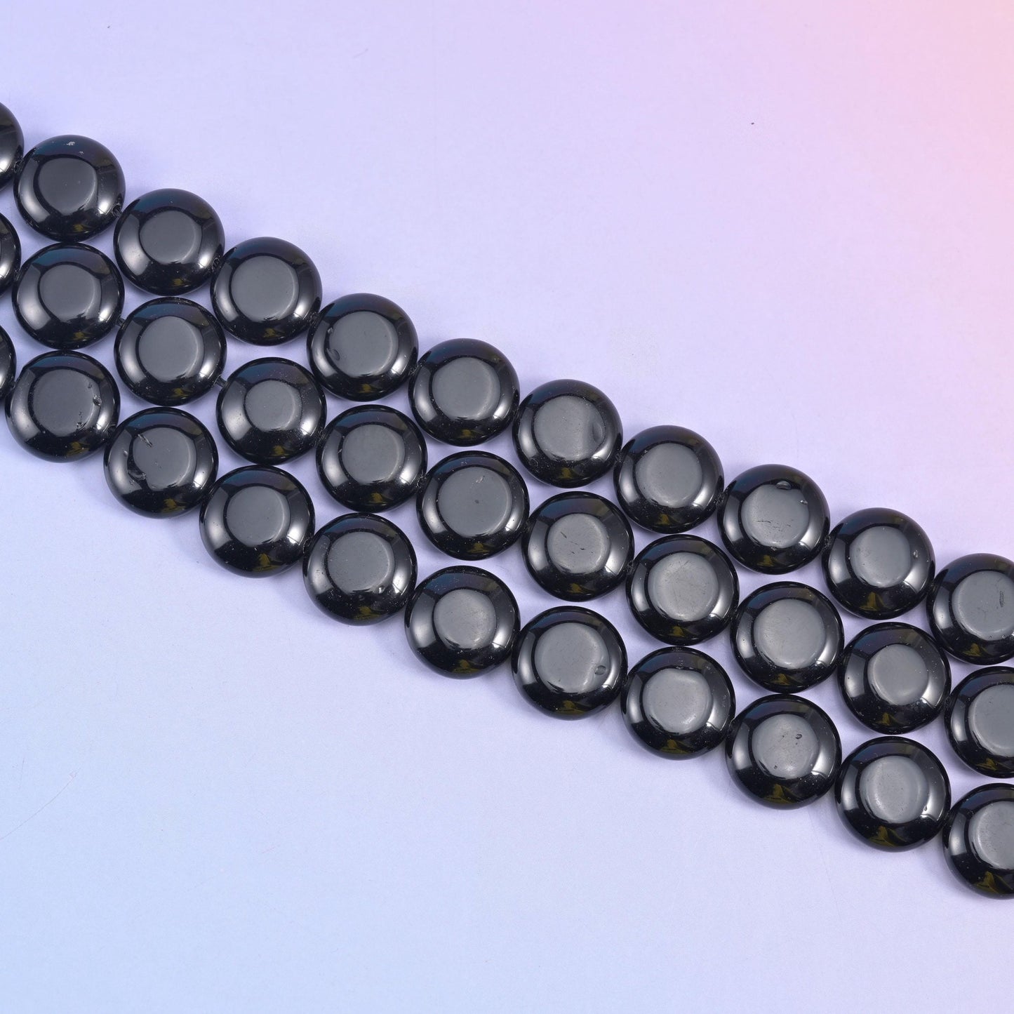 Diagonal rows of smooth obsidian coin beads on a pale lilac backdrop, each disc lying flat against the next
