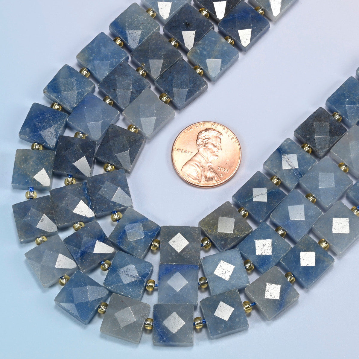 Close view of natural blue aventurine faceted flat square strands curving beside a US penny for scale