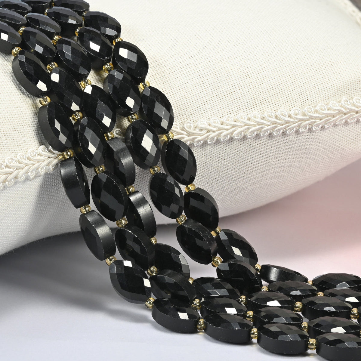 Black onyx faceted flat oval beads 9x14mm with small gold seed bead spacers, several strands draped over a white cushion