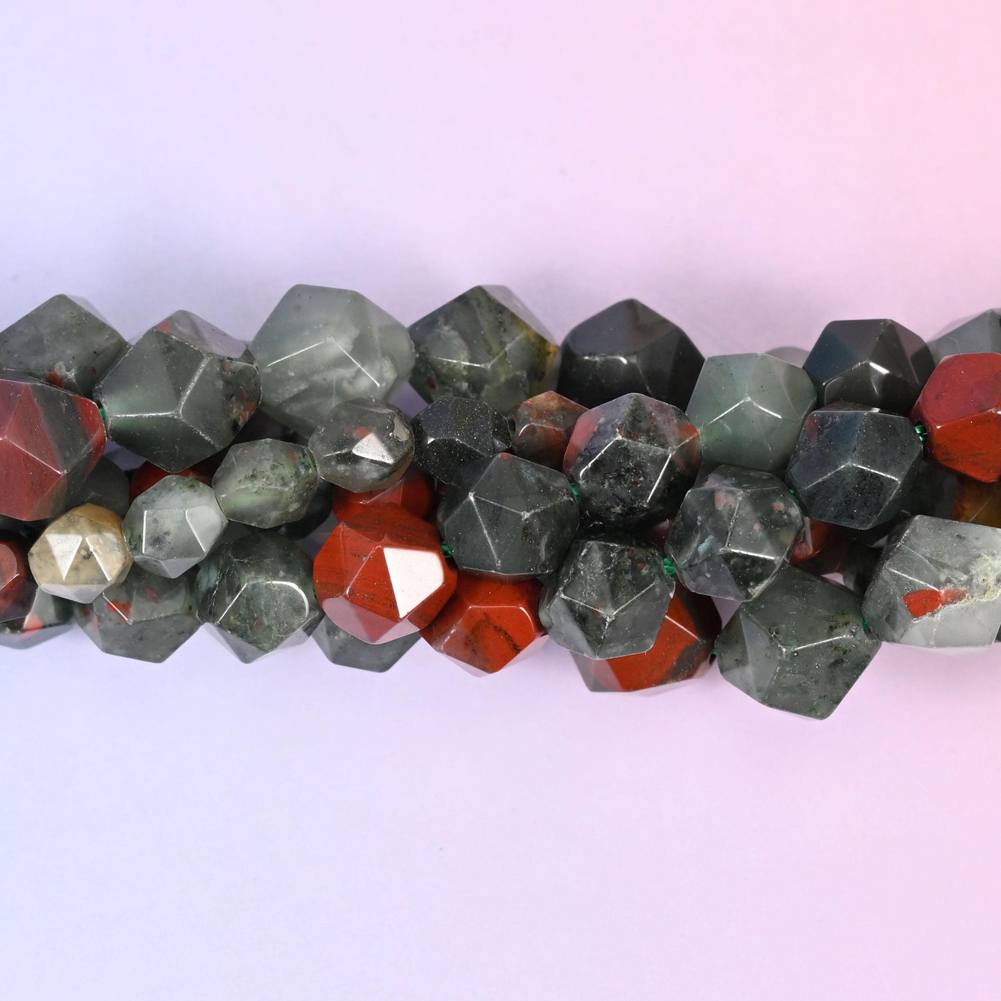 Sage and forest green beads splashed with brick red jasper, cut into faceted diamond stars so every flat plane picks up a hard little highlight. Some beads read nearly pale grey-green