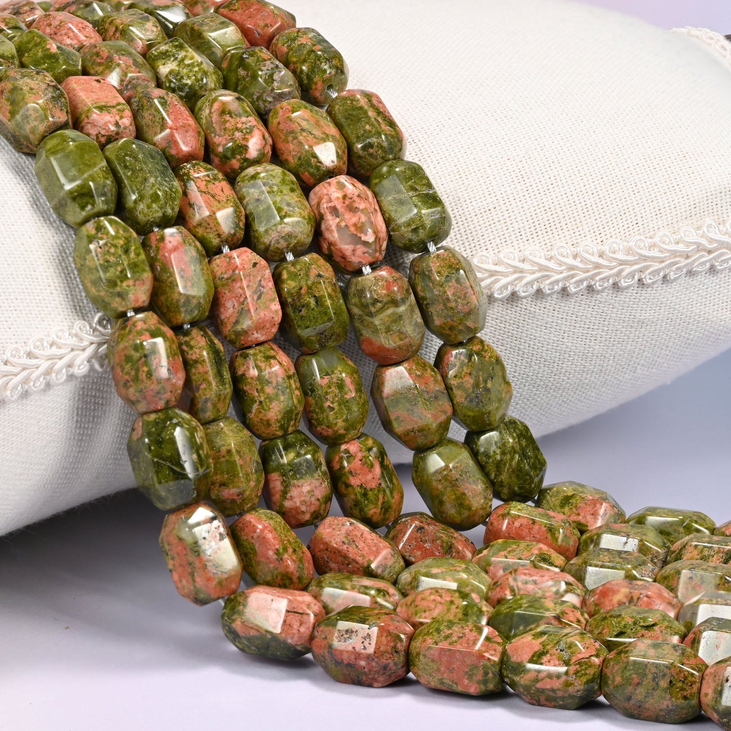 Close-up of multiple strands of green and salmon faceted Unakite barrel beads cascading diagonally over a cream woven pillow with decorative trim.