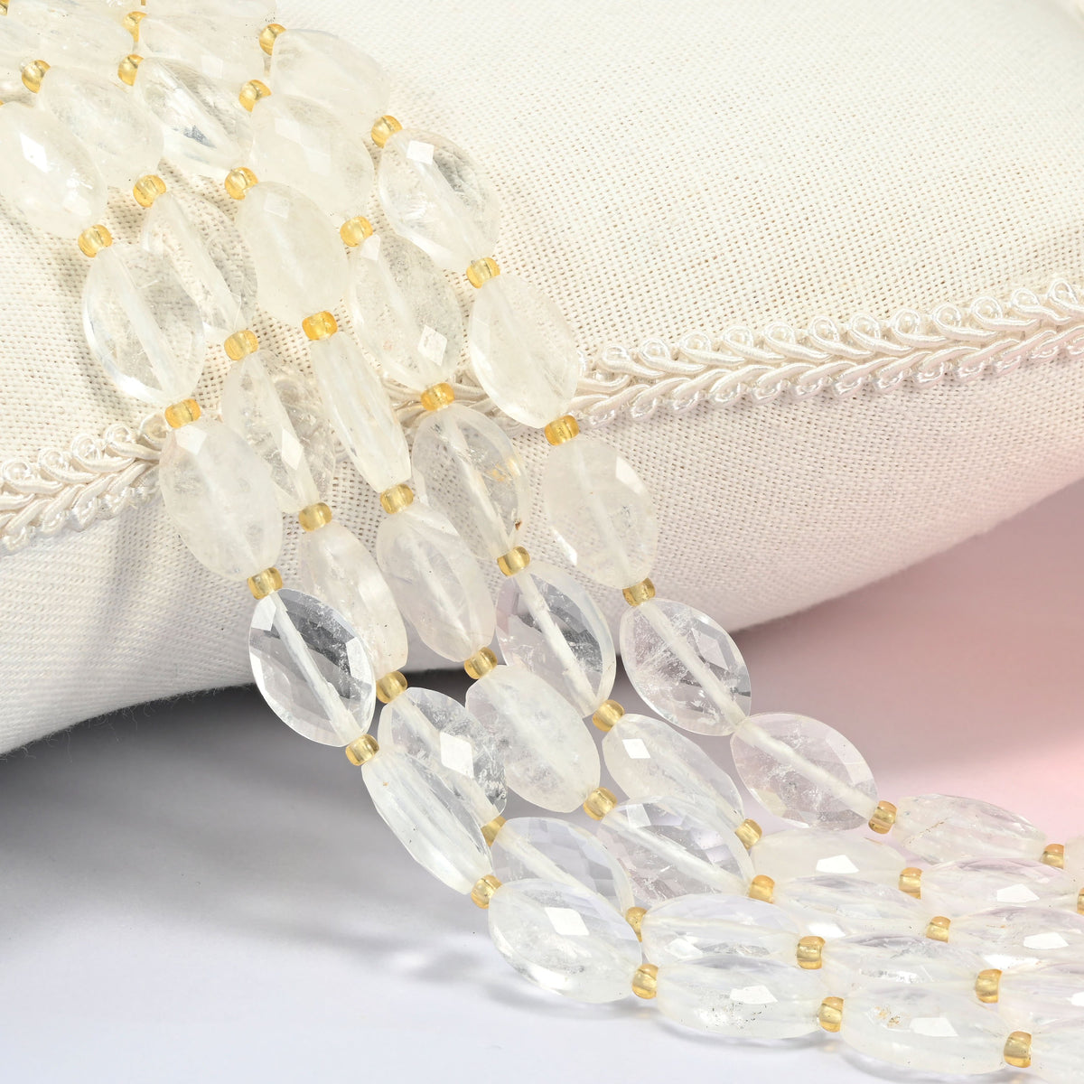 Clear quartz faceted flat oval beads 9x14mm with small gold seed bead spacers, several strands draped over a white cushion