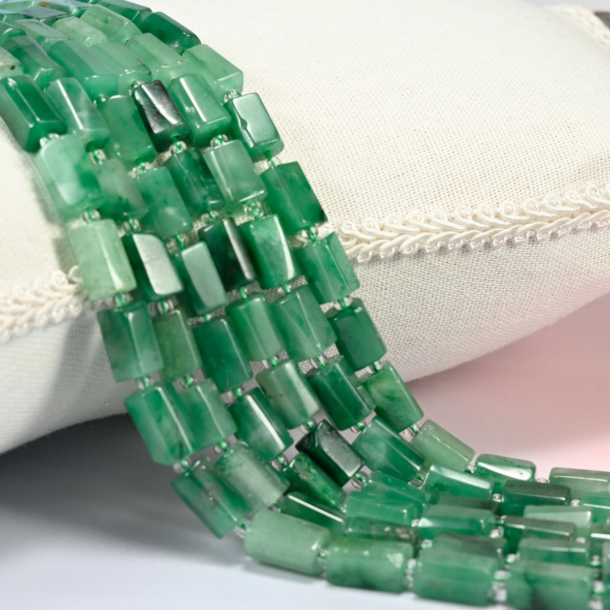 African jade faceted tube beads 7x11-7x12mm with small light green seed bead spacers, several strands draped over a white cushion