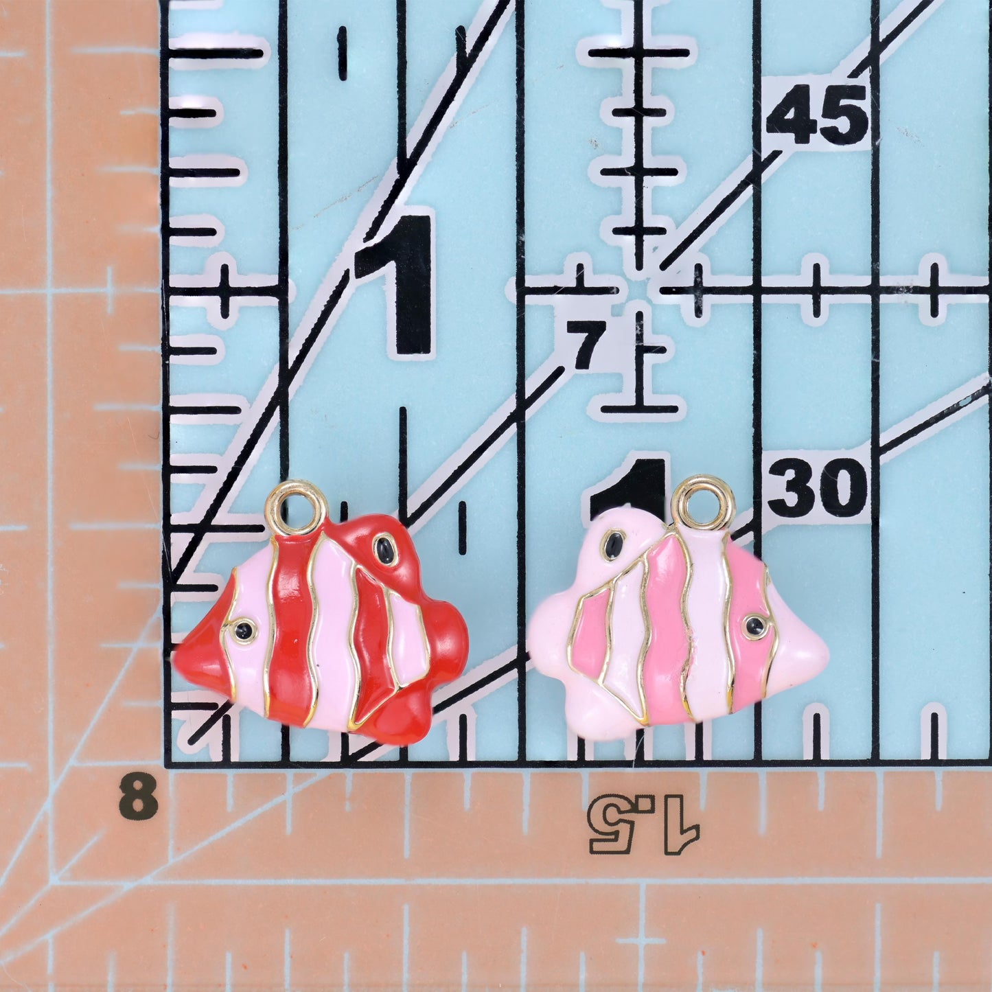 Enamel tropical fish charms in red and pink on a quilting ruler for size comparison.