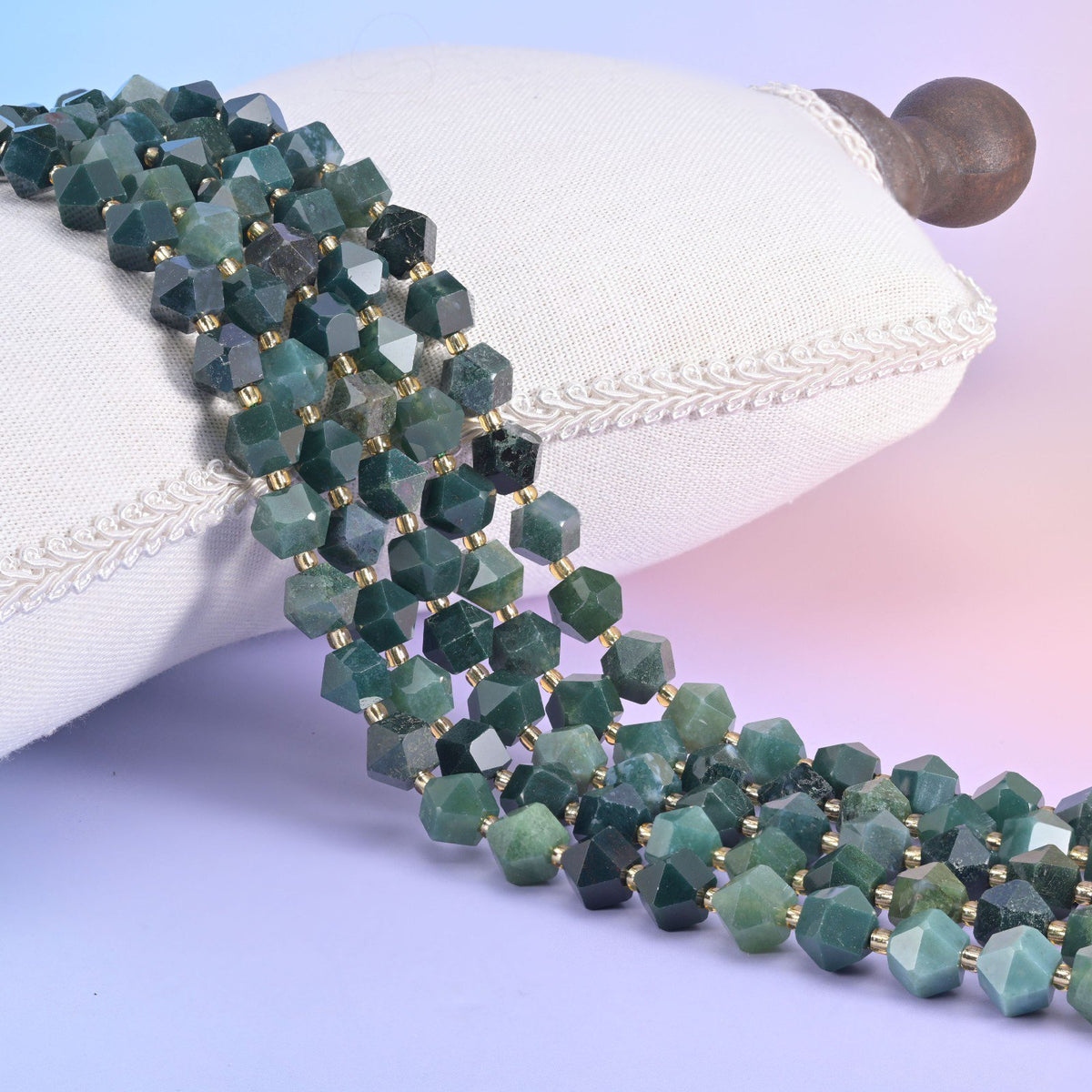 Faceted 8 sides diamond rhombus beads in moss agate draped over a white cushion, green moss patterning in clear stone.