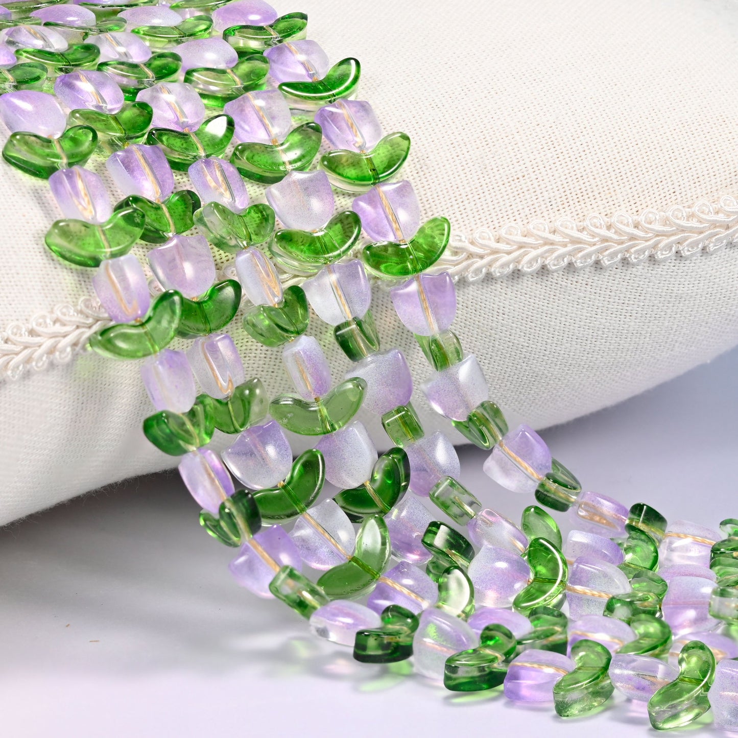 Close-up of lavender tulip flower and green leaf glass beads cascading diagonally over a cream woven pillow with decorative trim.
