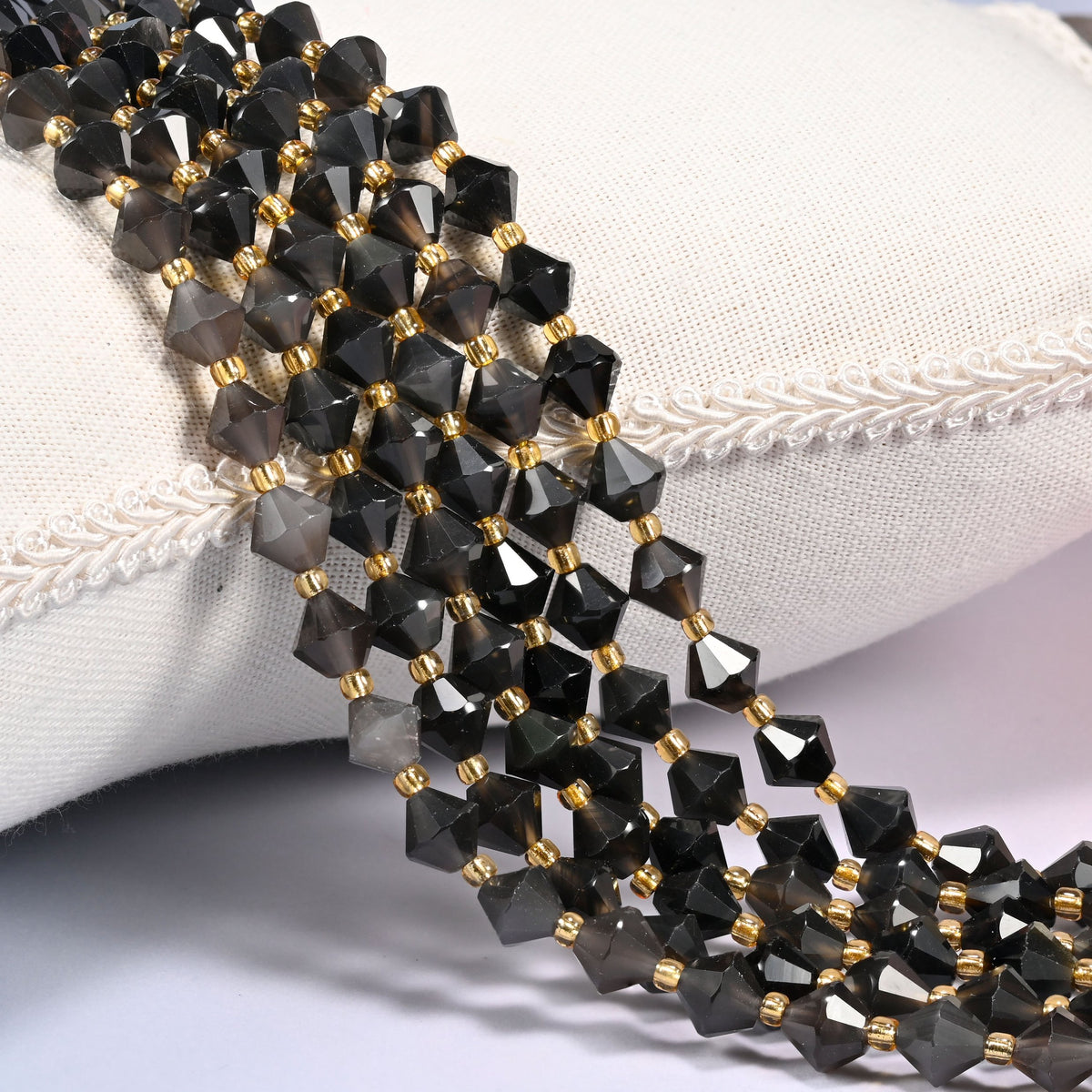 Ice Obsidian faceted bicone beads 8mm with gold-tone spacers, strand draped over a woven cushion trim