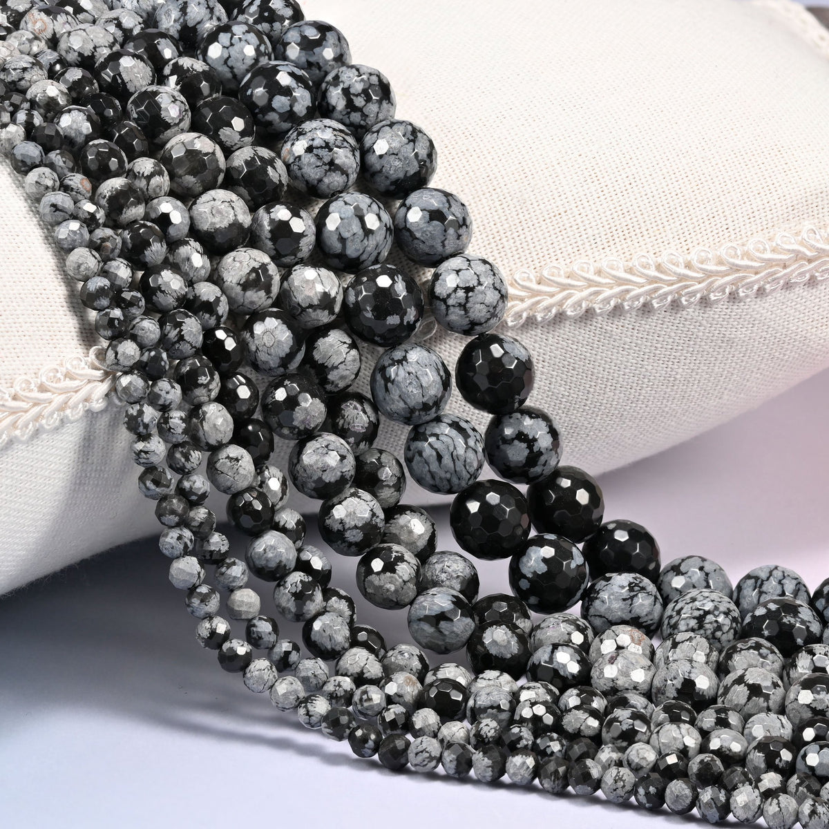Snowflake Jasper faceted round beads in graduating strand sizes coiled diagonally over a cream pillow with braided trim.