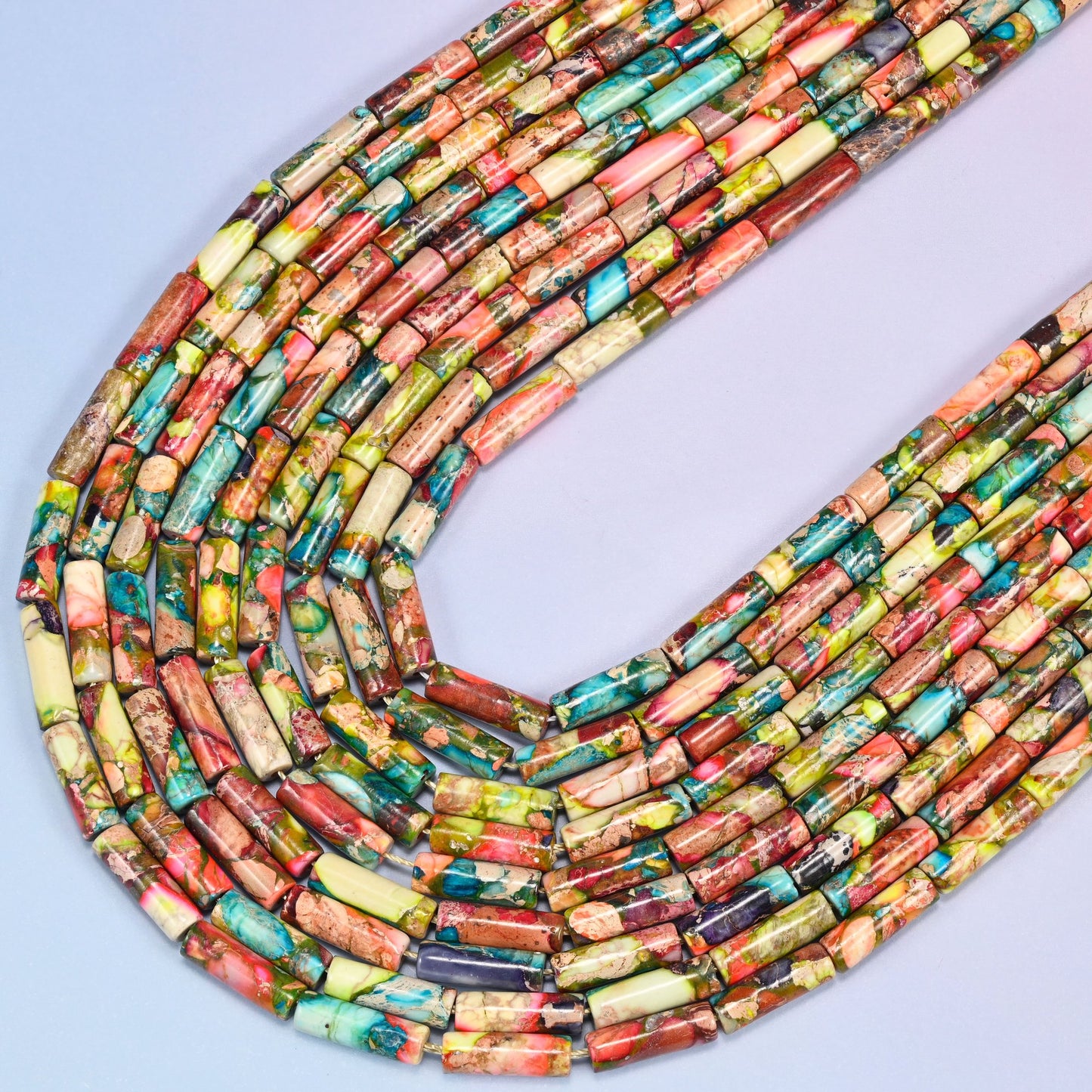 Close-up of colorful jasper tube beads coiled in a circular arrangement on a lavender background, showing turquoise, coral, and olive green coloring.