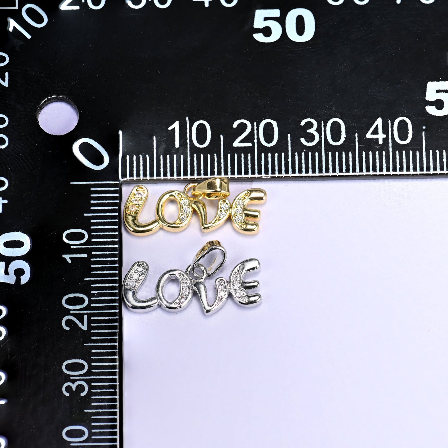 Gold and silver Love letter zirconia charms together on a black millimetre ruler, showing the script pendants at about 20mm wide.