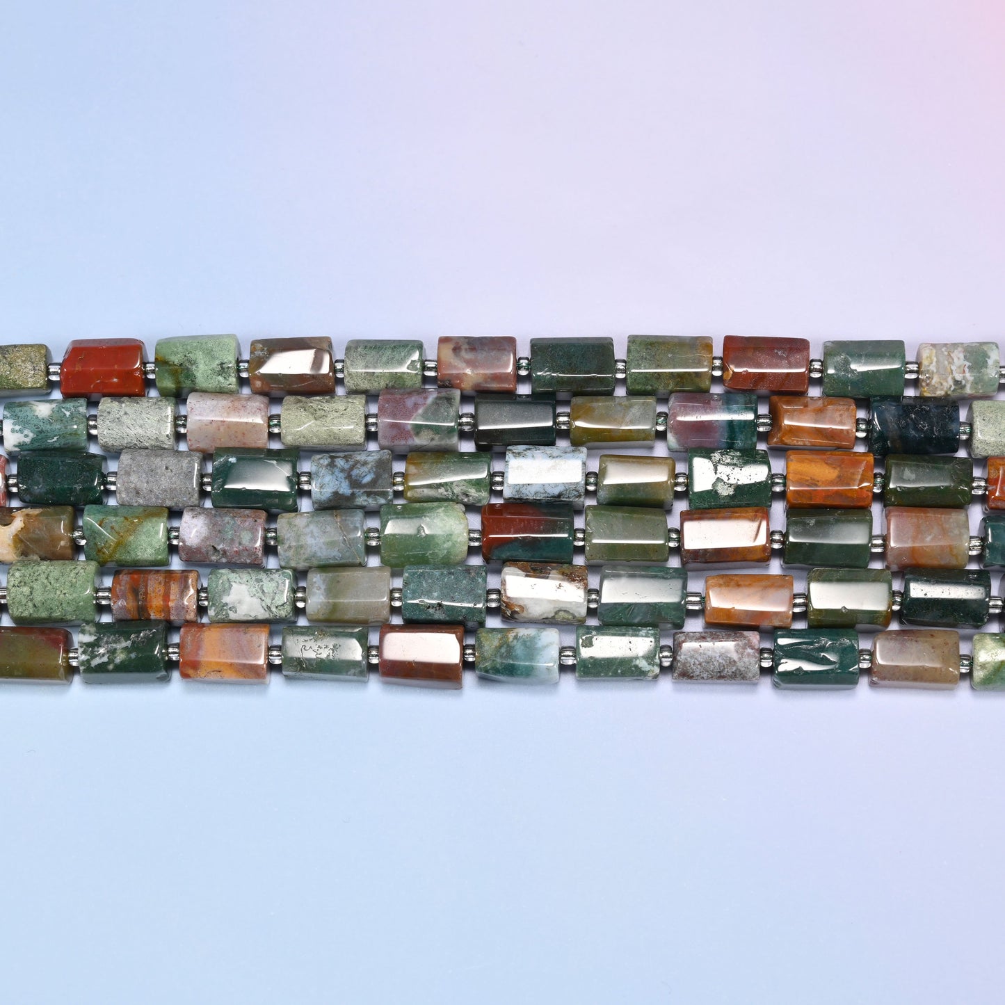 Overhead view of several rows of faceted Indian Agate tube beads in green, red, and cream tones on a light lavender surface.