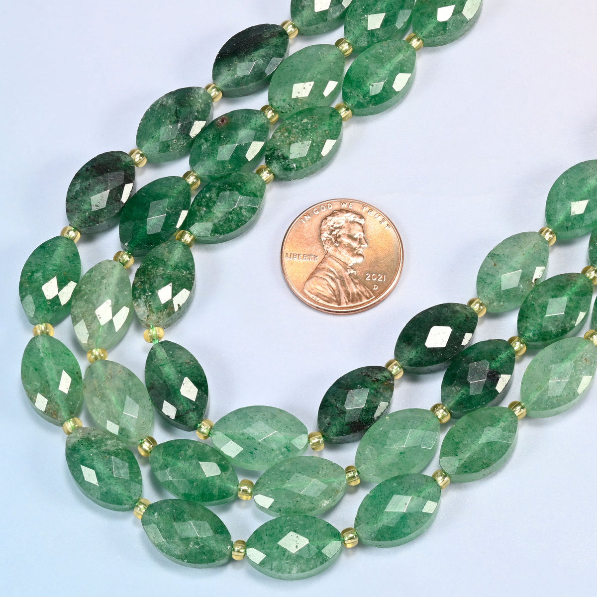 Close view of green strawberry quartz faceted flat oval strands curving beside a US penny for scale