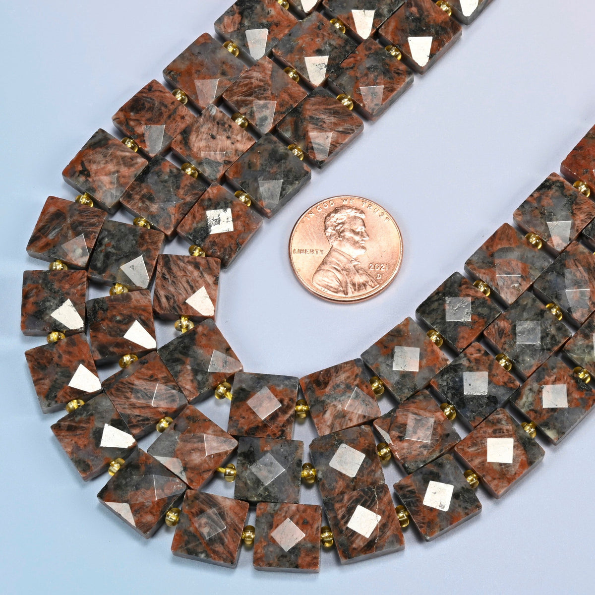 Close view of natural red sesame jasper faceted flat square strands curving beside a US penny for scale