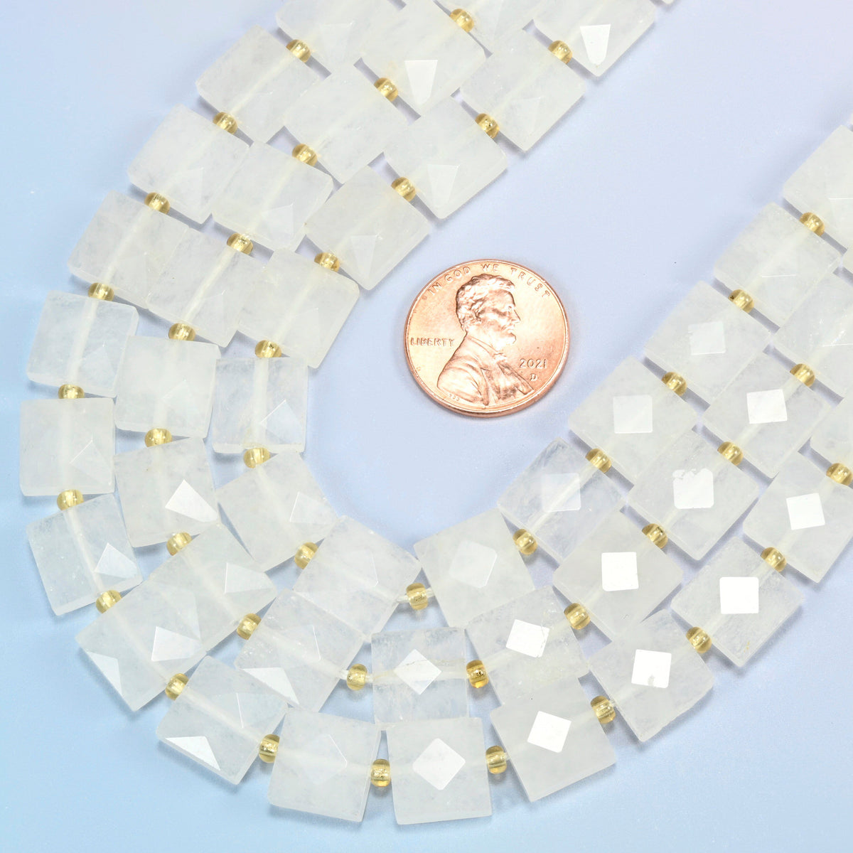 Close view of clear quartz faceted flat square strands curving beside a US penny for scale