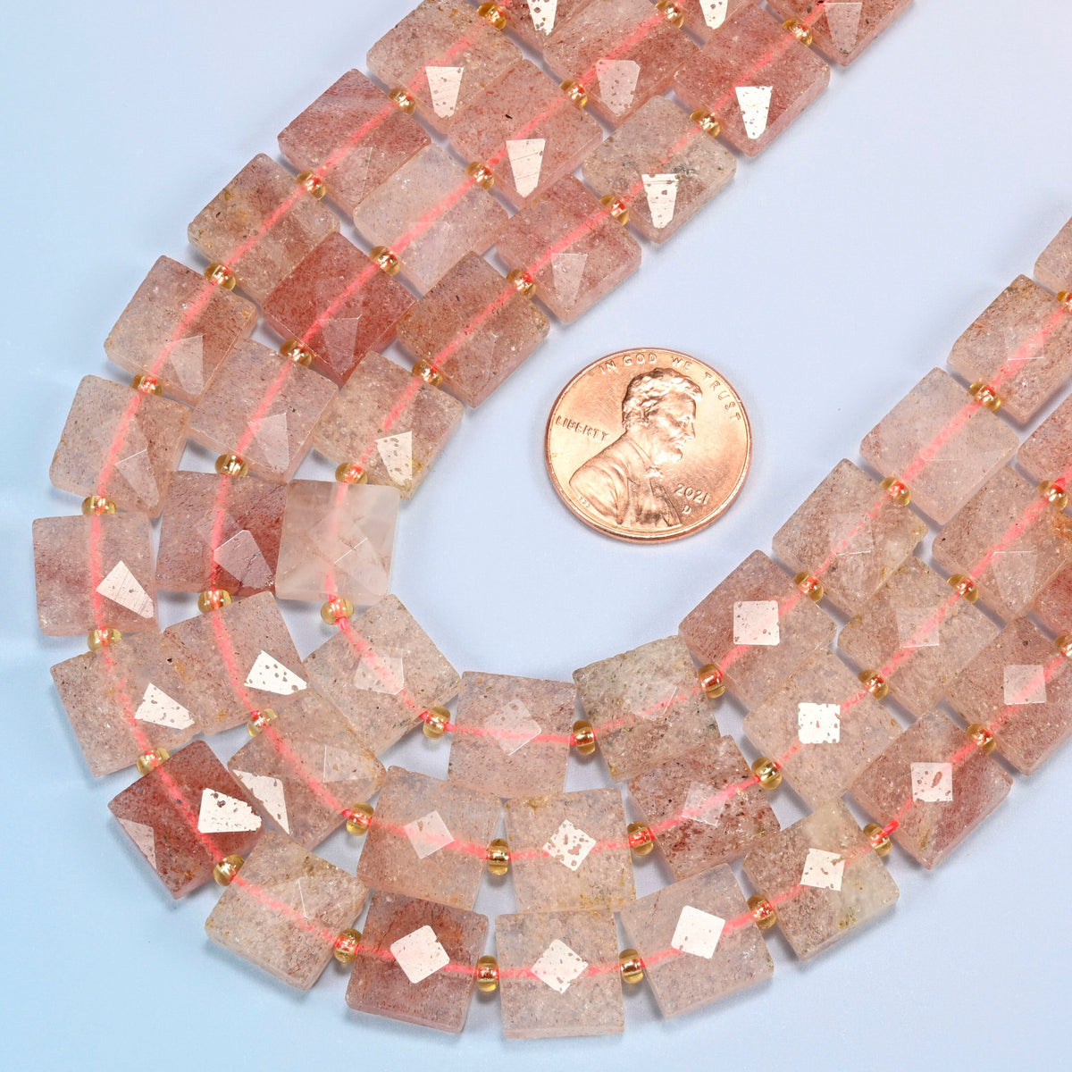 Close view of natural strawberry quartz faceted flat square strands curving beside a US penny for scale
